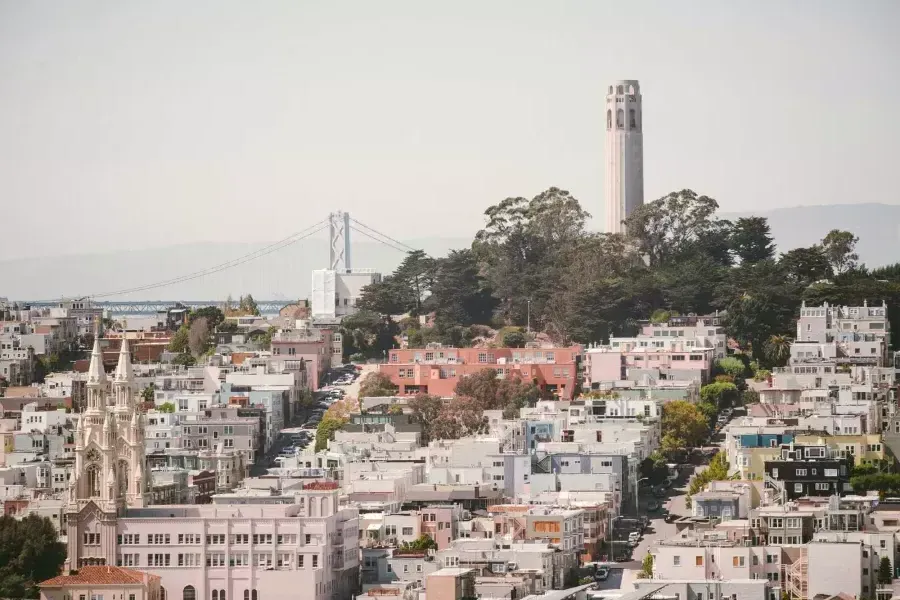 La Coit塔 di San Francisco è raffigurata con il Bay Bridge sullo sfondo e una collina ricoperta di case in primo piano.